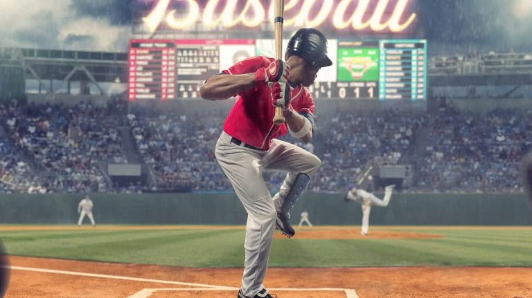 Baseball player in red jersey and gray pants swinging a bat on a baseball field with a crowd and scoreboard in the background.