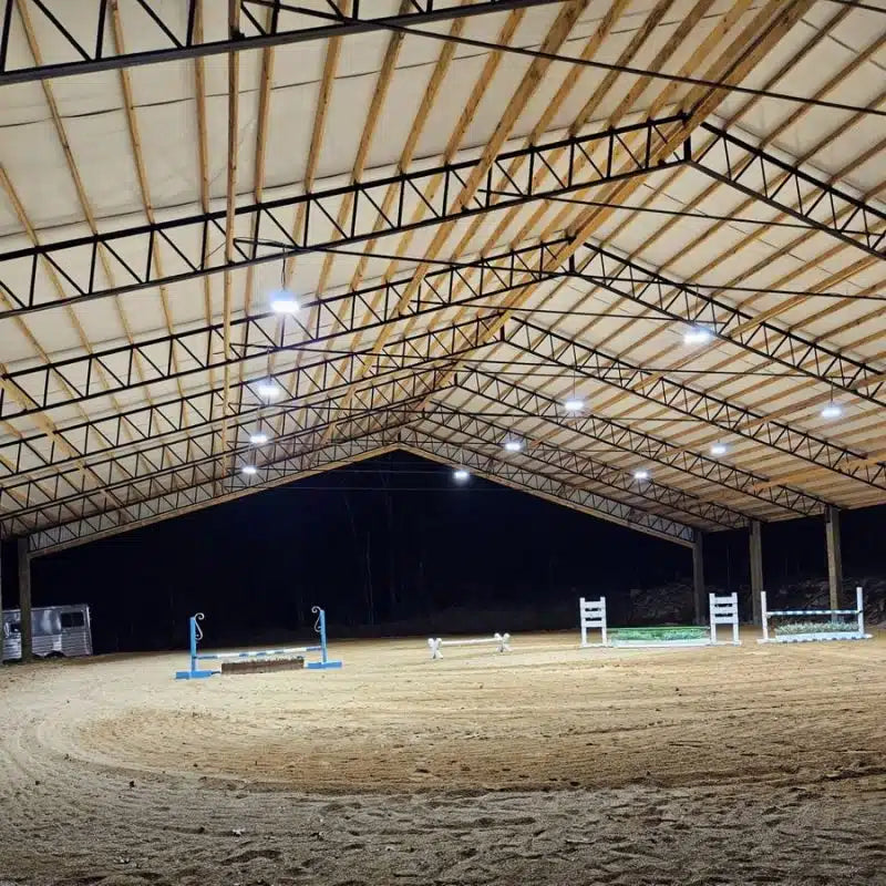 Interior of a large indoor riding arena with wooden roof beams and lighting fixtures.